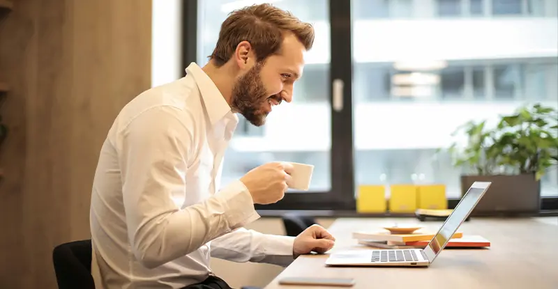 Man in white shirt smiling while working on laptop and holding a coffee cup in a modern office.